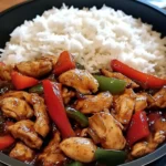 A sizzling garlic chicken stir-fry with colorful bell peppers served over steamed rice in a bowl, garnished with green onions and sesame seeds.